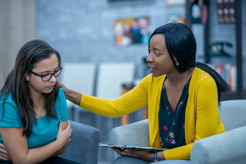 a black woman talking calmly to a woman that appears sad where the black woman is touching the woman on the shoulder