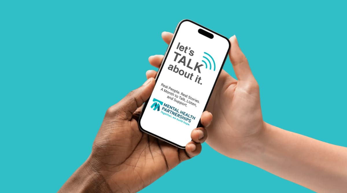 Two hands holding a mobile phone with the Let’s Talk About It campaign logo and slogan: “Real People. Real Stories. A Month to Talk, Listen, and Support.” The Mental Health Partnerships logo appears underneath the slogan.