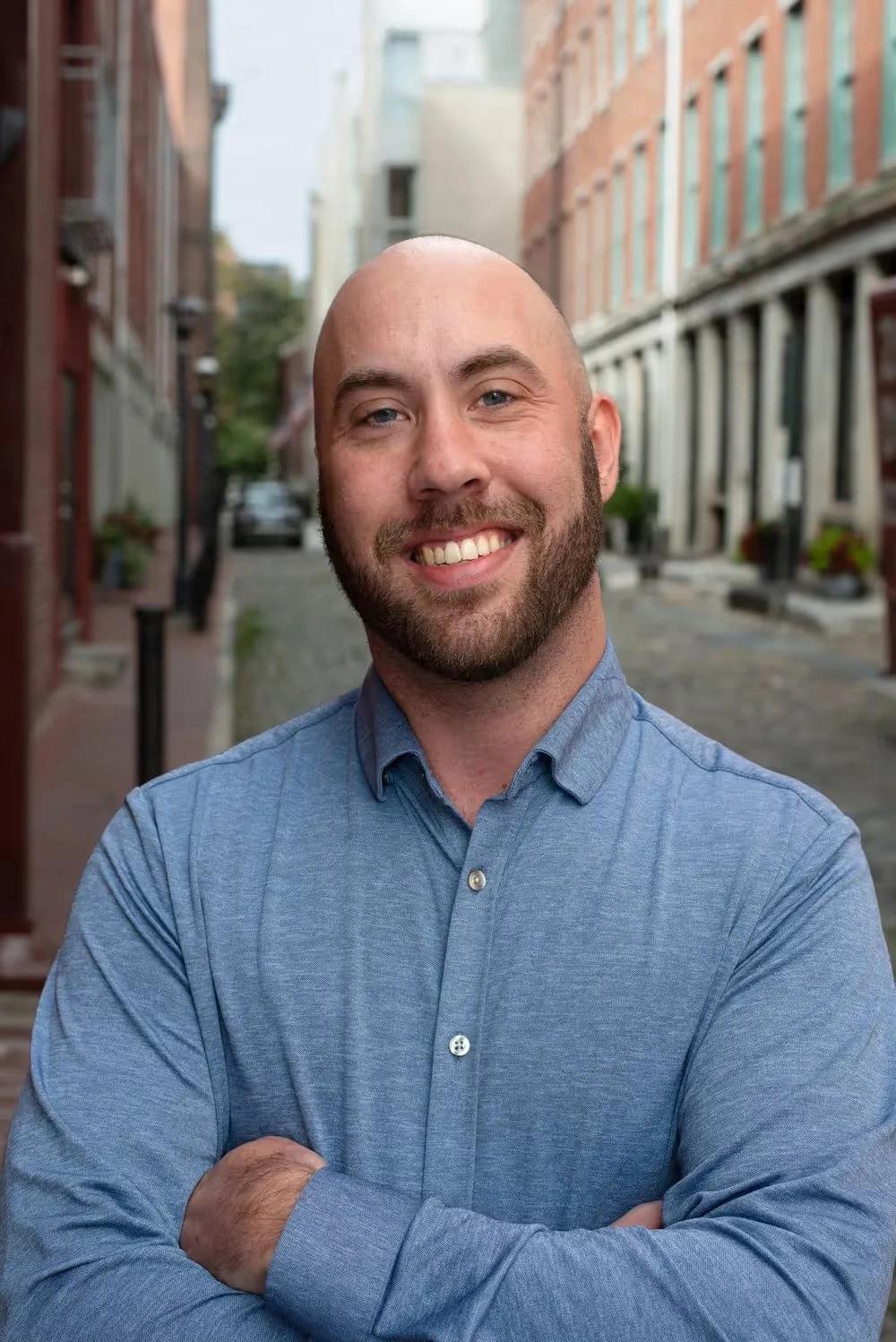 Jacob Bowling, a young white man with short light brown hair, smiling slightly while standing outside in a dark suit and tie against a brick wall. Board Member at Mental Health Partnerships.