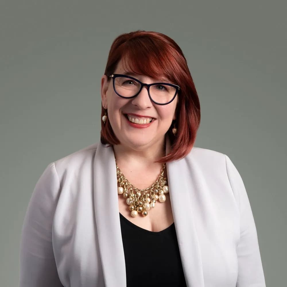 Leigh Braden, Division Director of The Institute at MHP, smiling and wearing glasses, a white blazer, and gold necklace.