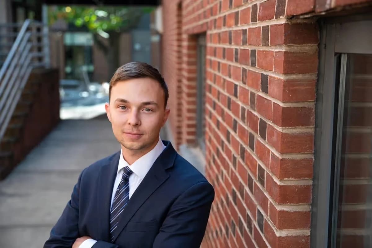 Josh Salzer, a white man with a shaved head and beard, smiling with arms crossed, wearing a light blue button-down shirt while standing in a cobblestone alleyway. Board Member at Mental Health Partnerships.