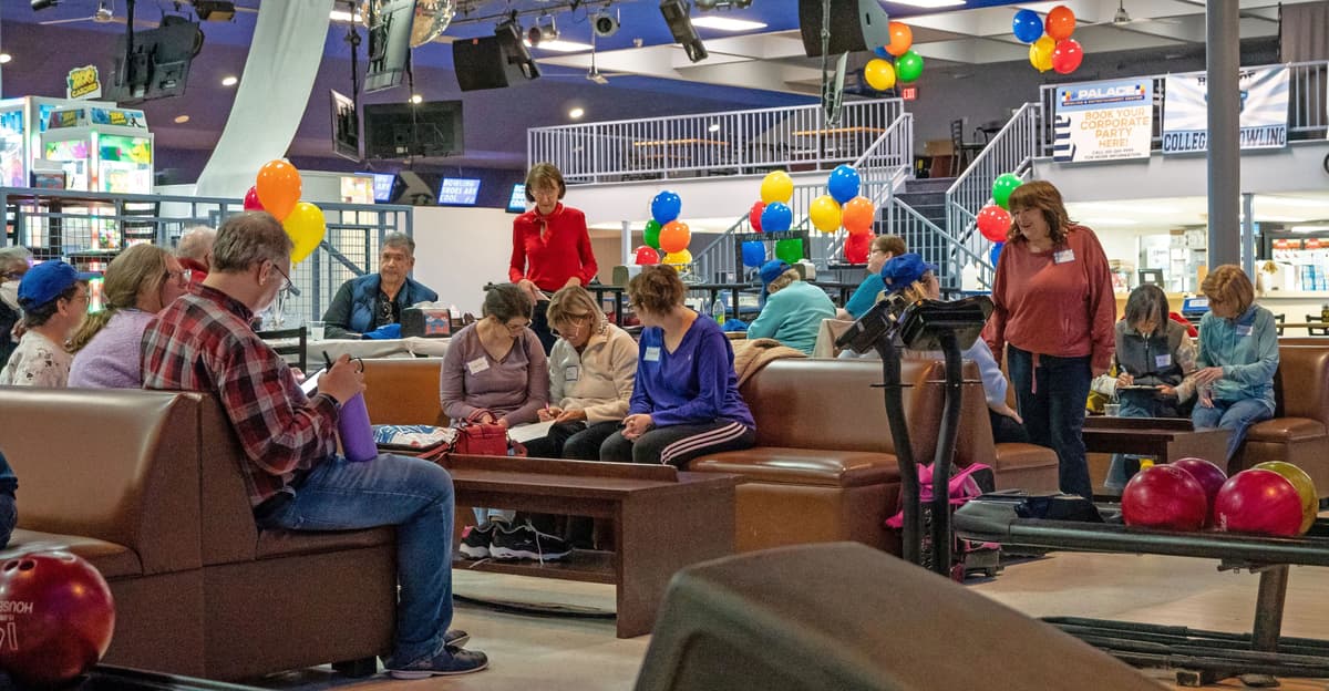A group of people playing bowling as part of an event. Balloons of varying colors in the background. 